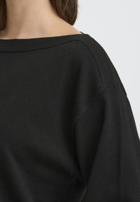 Žena v čiernom pletenom svetri so širokým výstrihom a spusteným ramenným švom, zobrazená od krku po hornú časť ramena.