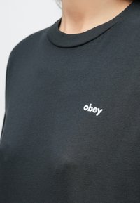 Person som har på seg en mørkegrå genser med rund hals og hvit skrift "obey" trykt på venstre bryst.