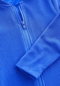 Blaue Zip-Jacke mit strukturiertem Oberflächenmaterial, geripptem Muster und langen Ärmeln. Reißverschlussdetail in der Mitte vorne sichtbar.