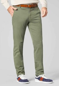 Olivgrüne Chinos mit slim fit, die eine glatte Textur aufweisen, vorne Taschen haben und mit einem braunen Ledergürtel kombiniert sind. Dazu weiße und marineblaue Sneakers.