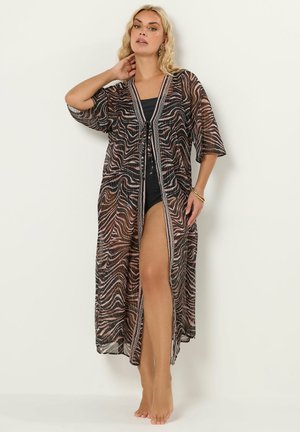 Lyshåret kvinde iført gennemsigtig zebra-stribet lang overdel med trekvartærme over sort badedragt, poserende barfodet mod en ensfarvet baggrund.