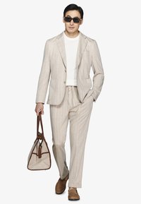 Uomo con completo beige a righe sottili, camicia bianca e occhiali da sole, che tiene una borsa a mano beige e marrone, camminando verso l'avanti.