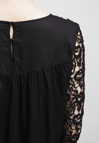 Sort top med rund halsudskæring, knaplukning bagpå og intrikate blonder detaljer på ærmerne. Blødt, flydende stof.