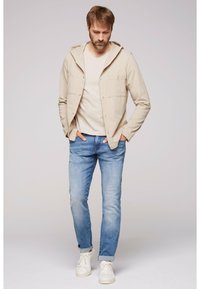 Chaqueta beige con capucha y dos bolsillos en el pecho, combinada con una camiseta beige clara y jeans de color azul claro, terminada con zapatillas blancas.