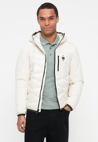 Veste blanche rembourrée avec capuche, dotée d'une fermeture éclair, d'une fermeture éclair noire contrastante et d'une petite poche sur la poitrine. Portée par-dessus un polo vert clair.