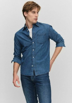 Denim shirt, dark blue, button-up design, single chest pocket, long sleeves rolled at cuffs, worn over a white T-shirt. Jeans in matching shade.