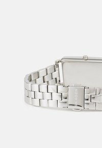 Silverfärgad klocka i rostfritt stål med rektangulärt hölje, länkarmbandsdesign och spännsklosur. Slät yta med minimal detaljering.