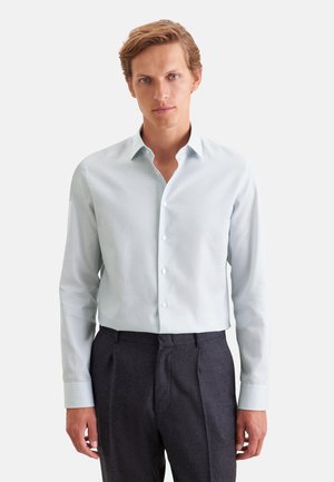 Homme aux cheveux châtain clair portant une chemise à manches longues claire à motifs et un pantalon formel sombre, debout devant un fond uni.