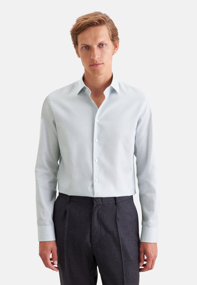 Homme aux cheveux châtain clair portant une chemise à manches longues claire à motifs et un pantalon formel sombre, debout devant un fond uni.