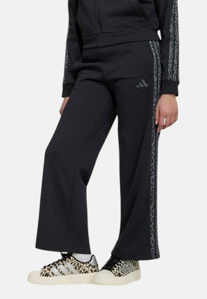 Mannequin portant un pantalon de survêtement Adidas noir évasé, avec des bandes latérales à motifs et des baskets à imprimé léopard.