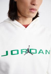 Fehér kapucnis pulóver textúrált anyagból, zöld "JORDAN" felirattal és fekete sziluett logóval az elején.