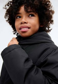 Enfant aux cheveux bouclés portant un manteau d'hiver noir, tirant sur le col, regardant vers le haut et souriant légèrement.