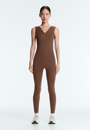 Kvinde stående iført en brun ærmeløs tætsiddende buksedragt med plisseret V-hals og hvide sneakers på en ensfarvet hvid baggrund.