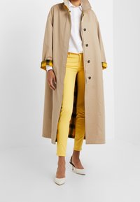 Abrigo largo beige con botones negros, forro de cuadros amarillos, llevado sobre una camisa blanca y pantalones amarillos. Tacones blancos completan el atuendo.