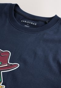 Tamsiai mėlynas medvilninis marškinėlis su apvalia iškirpte, priekyje spalvingas grafikas su skrybėle ir šventiniais akcentais. Matomas prekės ženklo ženklas.