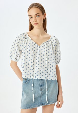 Witte blouse met blauwe geometrische patronen, V-hals, elastische pofmouwen en trekkoorddetail aan de onderkant, gecombineerd met een denim mini-jurk.
