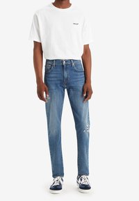 Bílý krátký rukáv T-shirt s logem "Levi's", kombinovaný se světle modrými džínami s poškozením na jednom koleni, nošený s navy teniskami.