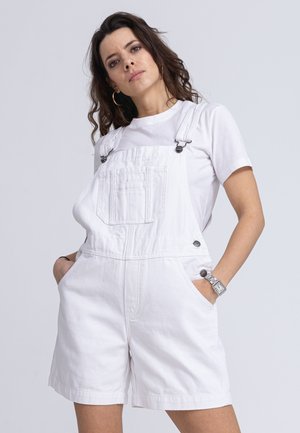 Weiße Jeanslatzhose mit verstellbaren Trägern, Fronttasche, Seitentasten und umgeschlagenen Säumen, getragen über einem weißen T-Shirt. Glatte Textur.