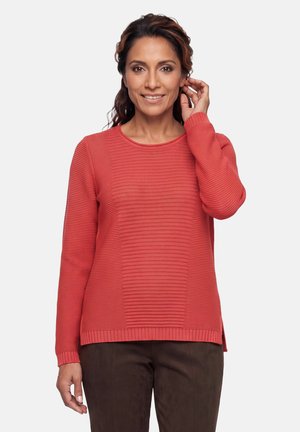 Woman with dark hair wearing a coral textured long-sleeve sweater and dark pants, smiling, with one hand touching her ear.