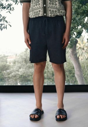 Person wearing black drawstring shorts, patterned short-sleeve shirt, and black woven slide sandals, standing indoors by a window with trees outside.