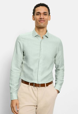 Mann trägt ein hellgrünes, langärmliges Hemd mit Knöpfen und beige Hosen mit braunem Gürtel, steht mit einer Hand in der Tasche.