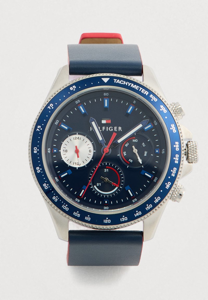 Orologio Tommy Hilfiger con quadrante blu, ghiera tachimetra, tre sottodial per 24 ore, giorno e data, e cinturino in pelle blu con parte inferiore rossa.