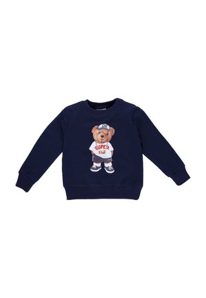 Marineblaue Sweatshirt mit einem Bärengrafik in einem weißen T-Shirt mit rotem "SUPER"-Druck, trägt graue Shorts und schwarze Sneaker.