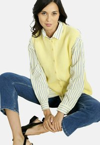 Geltona briaunuota liemenė virš mėlynos ir baltos juostuotos marškinėlių, derinama su trumpais džinsais ir juodais aukštakulniais sandalais. Modelis sėdi, rankos sukryžiuotos.