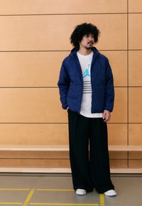 Mornarsko modra prešita jakna s kapuco preko bele majice s kratkimi rokavi, ki ima modro logotip. Črne široke hlače in bele čevlje z modrimi poudarki.