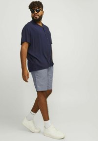 Chemise à manches courtes bleu marine avec boutons, shorts gris clair, baskets blanches et lunettes de soleil. Coupe moderne, tissu doux, look décontracté.