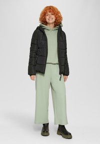 Schwarze Pufferjacke mit Kapuze über einem hellgrünen Hoodie und weit geschnittenen, knöchellangen Hosen. Schwarze klobige Stiefel runden das Outfit ab.