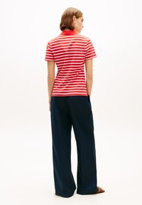 Γυναίκα στέκεται με την πλάτη, φοράει κόκκινη και λευκή ριγωτή κοντομάνικη μπλούζα, σκούρο μπλε παντελόνι με φαρδιά μπατζάκια και καφέ σανδάλια.