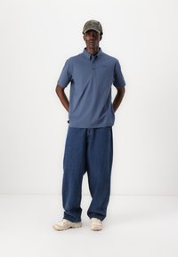 Polo majica iz modrega blaga s ovratnikom in logotipom, v kombinaciji z ohlapnimi denim hlačami in bež športnimi copati, dopolnjena z kapo z vzorcem.