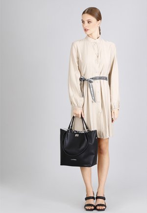 Beige knopenjurk met een zwart-wit patroonriem, gecombineerd met een zwarte leren totebag met accenthandvatten en een logo.