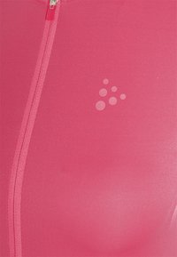 Omezený růžový tílko s předním zipem, hladkou texturou a diskrétním logem s různě velkými kruhy na levé straně.