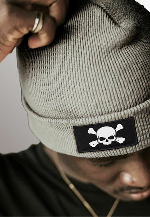 Gray knitted beanie with a black rectangular patch featuring a white skull and crossed bones. The texture is ribbed, providing a snug fit.