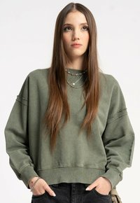 Młoda kobieta z długimi brązowymi włosami, ubrana w za dużą oliwkową bluzę i warstwowe srebrne naszyjniki, z rękami schowanymi w kieszeniach czarnych spodni.