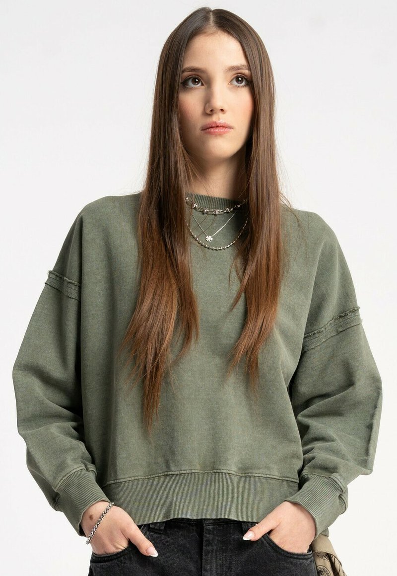 Młoda kobieta z długimi brązowymi włosami, ubrana w za dużą oliwkową bluzę i warstwowe srebrne naszyjniki, z rękami schowanymi w kieszeniach czarnych spodni.