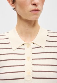 Πολό πουκάμισο σε κρέμα απόχρωση με ριγέ υφή, που διαθέτει αιχμηρό γιακά και τέσσερα κρέμα κουμπιά στη ρίγα. Λεπτομέρειες σε καφέ ρίγες.