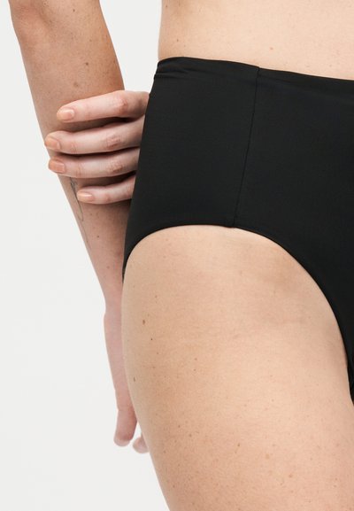 Κοντινό σε γοφό και χέρι ατόμου που φοράει μαύρο μαγιό με ψηλή μέση, με απλό ανοιχτό φόντο.