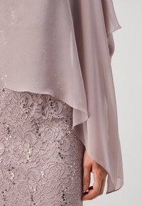 Main reposant sur le côté, portant une manche en chiffon mauve sur une jupe en dentelle avec des embellissements en sequins.
