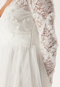 Robe blanche avec un corsage en dentelle orné d'applications florales, manches longues transparentes et jupe en tulle fluide. Détail de fermeture éclair au dos.