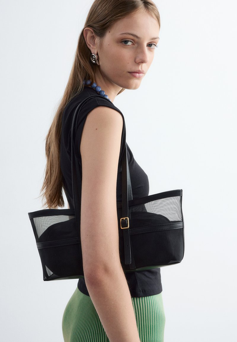 Jeune femme aux longs cheveux bruns porte un haut noir sans manches, un pantalon vert côtelé et un sac à bandoulière noir en maille.