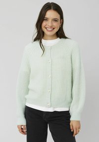 Cardigan en tricot vert clair avec un motif texturé, col rond et cinq boutons blancs sur le devant. Porté sur une chemise blanche.