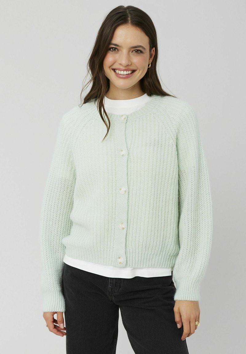 Cardigan en tricot vert clair avec un motif texturé, col rond et cinq boutons blancs sur le devant. Porté sur une chemise blanche.