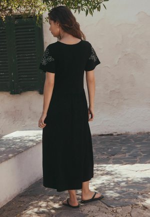 Žena stojící venku na kamenné dlažbě, má na sobě černé šaty délky ke kotníkům s květinovou výšivkou na rukávech a černé sandály.