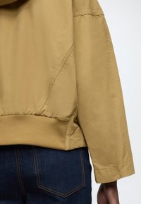 Keki színű, sima anyagból készült dzseki, bordázott szegéllyel és mandzsettákkal, kényelmes szabással és finom oldalsó varrási részletekkel. Sötétkék farmernadrág látható.