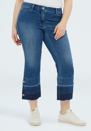 Vaqueros azules de corte medio y largo hasta el tobillo con detalles de botones y dobladillos sin rematar, combinados con zapatillas blancas y una camiseta blanca.