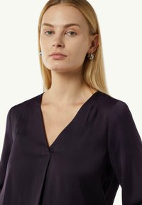 Dunkelviolette Bluse mit V-Ausschnitt, aus glattem Material, mit dezenten Falten am Ausschnitt und langen Ärmeln. Silberne Creolen sind sichtbar.
