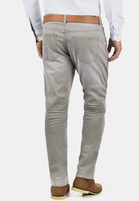 Grå slim-fit jeans med en glatt tekstur, fem lommer og et brunt lærbelte, sett bakfra. Støvler med en lys såle.